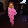 child, toddler, girl, portrait, smiling, standing, pink_clothing, tracksuit, sweatshirt, pants, braids, indoors, living_room, furniture, armchair, window, curtains, carpet, barefoot, fashion