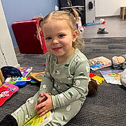 Harper is registered to the contest to win money with this photo: toddler, child, pigtails, book, carpet, toys, indoor, smiling, clothing, floor, reading, playroom, young_child, person, cute, happy, casual, furniture, background, portrait
