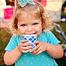 toddler, child, girl, blue_eyes, bow, turquoise_dress, polka_dots, paper_cup, smiling, happy, outdoor, portrait, curly_hair, cute, hands, face, person, daylight, background_blur, playful