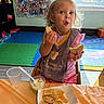 toddler, child, snack, tortilla_chips, bib, table, plastic_cup, indoor, playroom, colorful, wall_collage, high_chair, food, happy, messy_face, curly_hair, pink_clothing, floor_mat, window, daylight