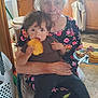 child, elderly_person, bread, kitchen, food, floral_dress, curly_hair, seated, plate, fork, cabinet, floor, laundry_basket, toddler, person, home_interior, snack, holding, meal, casual_clothing