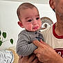 Callan is registered to the contest to win money with this photo: baby, crying, adult_hand, indoor, person, holding, pout, gray_clothing, face, expression, tears, skin, head, plant, couch, blanket, basket, home, comfort, closeup