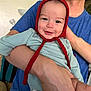 baby, infant, smile, red_bonnet, blue_shirt, adult_arm, child, happy, portrait, indoors, person, cute, holding, face, clothing, skin, hand, headwear, expression, warmth