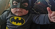Oliver joined the competition — help win amazing prizes! baby, batman_costume, thumbs_up, child, mask, superhero, indoor, couch, blanket, television, person, costume, cute, smile, seat, home, furniture, childhood, fun, portrait