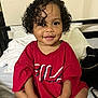 Nayla joined the competition — help win amazing prizes! toddler, child, smiling, curly_hair, red_tshirt, sitting, bed, pillow, indoors, person, cute, happy, portrait, clothing, dimple, young_child, casual, cozy, room, furniture
