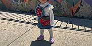Nayla is registered to the contest to win money with this photo: toddler, child, curly_hair, denim_dress, strawberry_design, purple_crocs, sidewalk, shadow, playground, fence, signs, colorful_wall, sunlight, outdoor, urban, young_child, concrete, daytime, curious_expression, standing