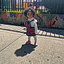 toddler, child, curly_hair, denim_dress, strawberry_design, purple_crocs, sidewalk, shadow, playground, fence, signs, colorful_wall, sunlight, outdoor, urban, young_child, concrete, daytime, curious_expression, standing