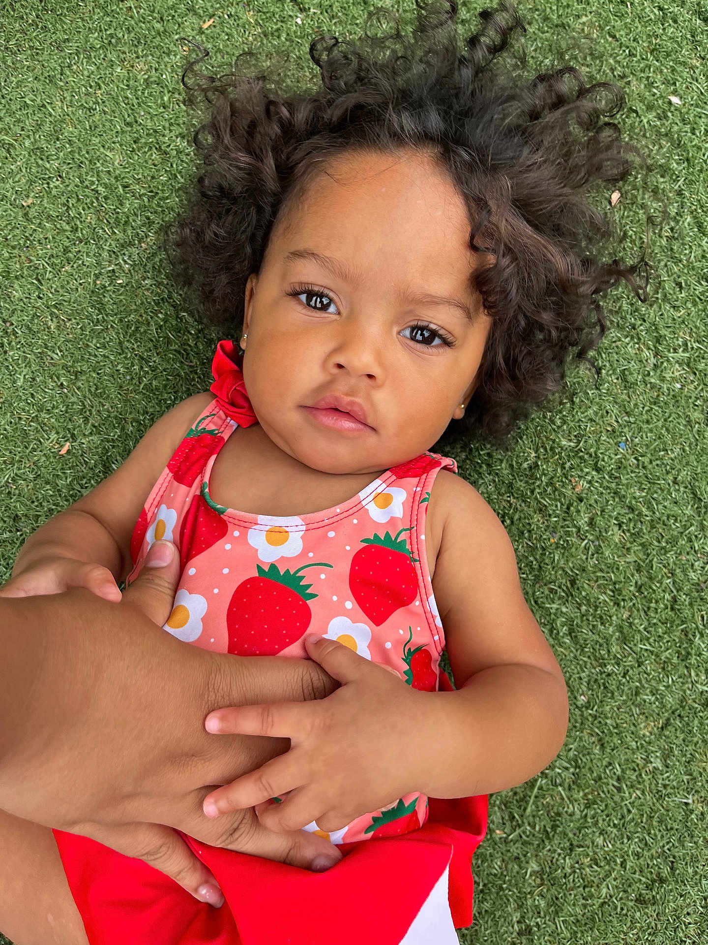Nayla is registered to the contest to win money with this photo: toddler, child, curly_hair, grass, outdoor, hand, holding, strawberry_pattern, red_clothing, cute, portrait, young_child, person, skin, closeup, baby, summer, nature, casual, expression