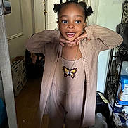 Blue is registered to the contest to win money with this photo: child, girl, smiling, posing, indoors, long_sleeve, cardigan, jumpsuit, butterfly_necklace, hair_buns, curly_hair, wooden_floor, door, household_items, storage_shelf, water_jug, casual_clothing, happy, portrait, young