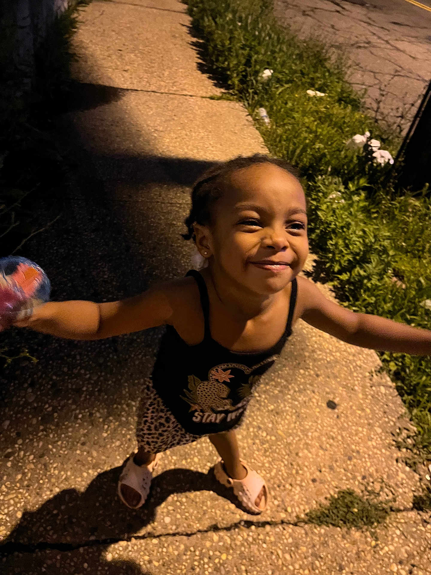 Blue joined the competition — help win amazing prizes! child, girl, smiling, sidewalk, night, braids, happy, streetlight, shadow, plants, grass, concrete, footwear, sleeveless_top, patterned_skirt, outdoors, playful, holding_object, person, casual