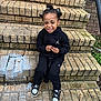 child, smiling, sitting, brick_steps, black_hoodie, black_pants, sneakers, outdoor, playful, person, hair_buns, grin, concrete, casual_clothing, young_child, urban, daylight, holding_object, texture, greenery