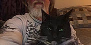 Sneezer is registered to the contest to win money with this photo: man, cat, indoor, glasses, beard, black_cat, white_paws, couch, curtains, casual_clothing, portrait, pet, relaxed, seated, home, feline, whiskers, green_eyes, facial_hair, long_hair