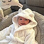 baby, smile, hooded_robe, white_clothing, plush_toy, cloud_toy, cushion, indoor, cozy, soft_texture, child, infant, happy, face, person, cute, seated, pink_blanket, furniture, relaxing