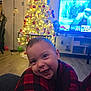 Bresman Jr is registered to the contest to win money with this photo: baby, child, smiling, face, plaid, pajamas, knee, christmas_tree, lights, living_room, television, floor, furniture, decorations, holiday, indoors, happy, person, wood_floor, cozy