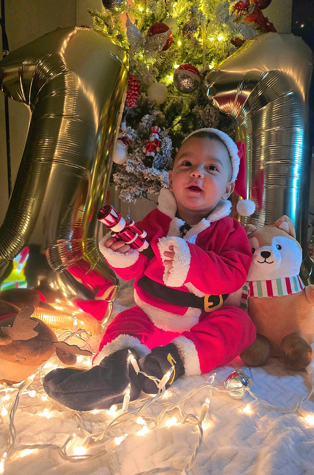 Bresman Jr is registered to the contest to win money with this photo: baby, child, christmas_tree, holiday_lights, santa_claus_outfit, plush_toy, balloon, number_balloons, festive, indoors, portrait, smiling, celebration, holiday_decor, toy, red_clothing, white_fur, black_boots, face, festive_hat