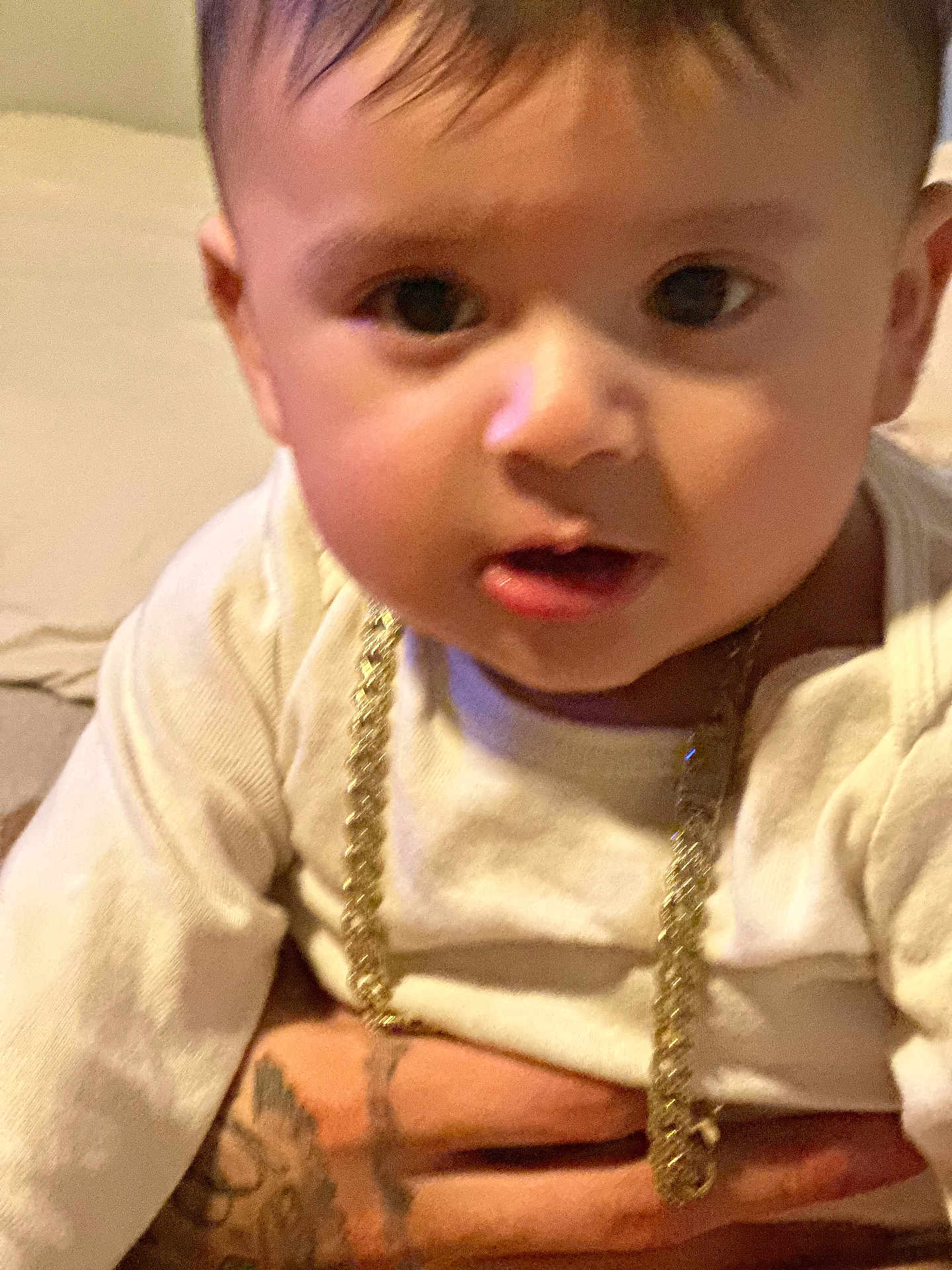Walter is registered to the contest to win money with this photo: baby, child, face, gold_chain, white_clothing, tattoo, hand, curious, portrait, closeup, indoor, skin, person, cute, infant, expression, holding, young, head, human
