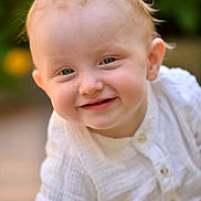Mason is registered to the contest to win money with this photo: baby, child, smile, face, blue_eyes, light_hair, white_shirt, portrait, outdoor, garden, flower, blurred_background, happy, cute, infant, close_up, person, skin, head, childhood