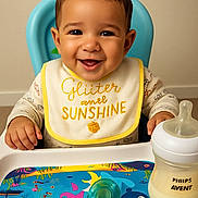 Sidi Baba is registered to the contest to win money with this photo: baby, smiling, high_chair, bib, milk_bottle, pacifier, colorful_placemat, indoor, child, cute, happy, feeding_time, infant, face, person, young_child, table, toy, baby_food, nursery