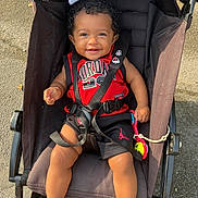 Kayla participe au concours pour gagner de l'argent avec cette photo : baby, child, stroller, smiling, curly_hair, red_clothing, black_clothing, outdoor, sunlight, happy, person, seat_belt, toy, pavement, casual, summer, baby_clothes, legs, hands, face