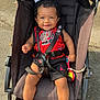 baby, child, stroller, smiling, curly_hair, red_clothing, black_clothing, outdoor, sunlight, happy, person, seat_belt, toy, pavement, casual, summer, baby_clothes, legs, hands, face