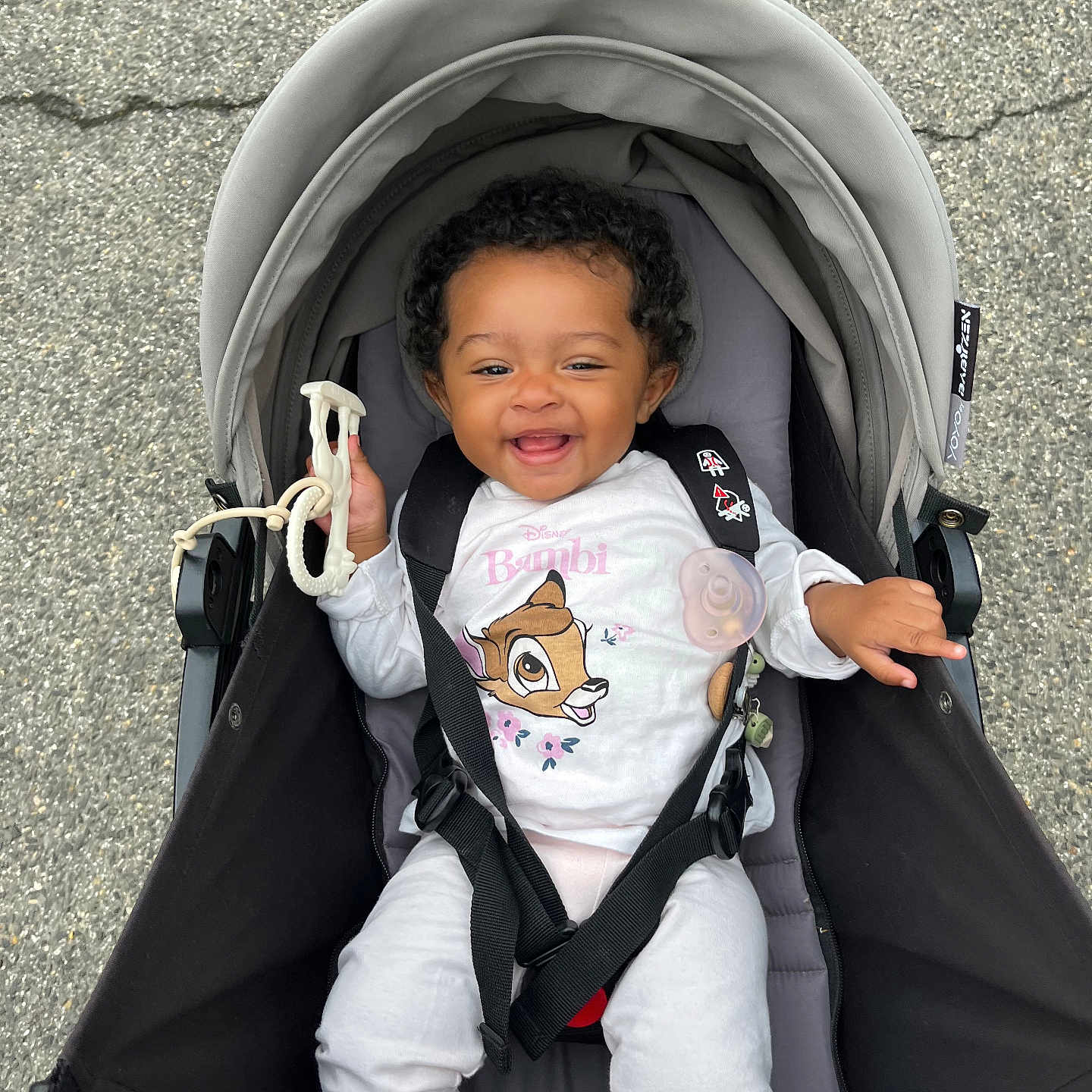 Kayla a rejoint le concours — aidez-le/la à gagner de superbes lots ! baby, bambi, black_straps, child, clothing, curly_hair, cute, daylight, happy, hood, infant, outdoor, pavement, person, playful, seatbelt, sitting, smiling, stroller, toy