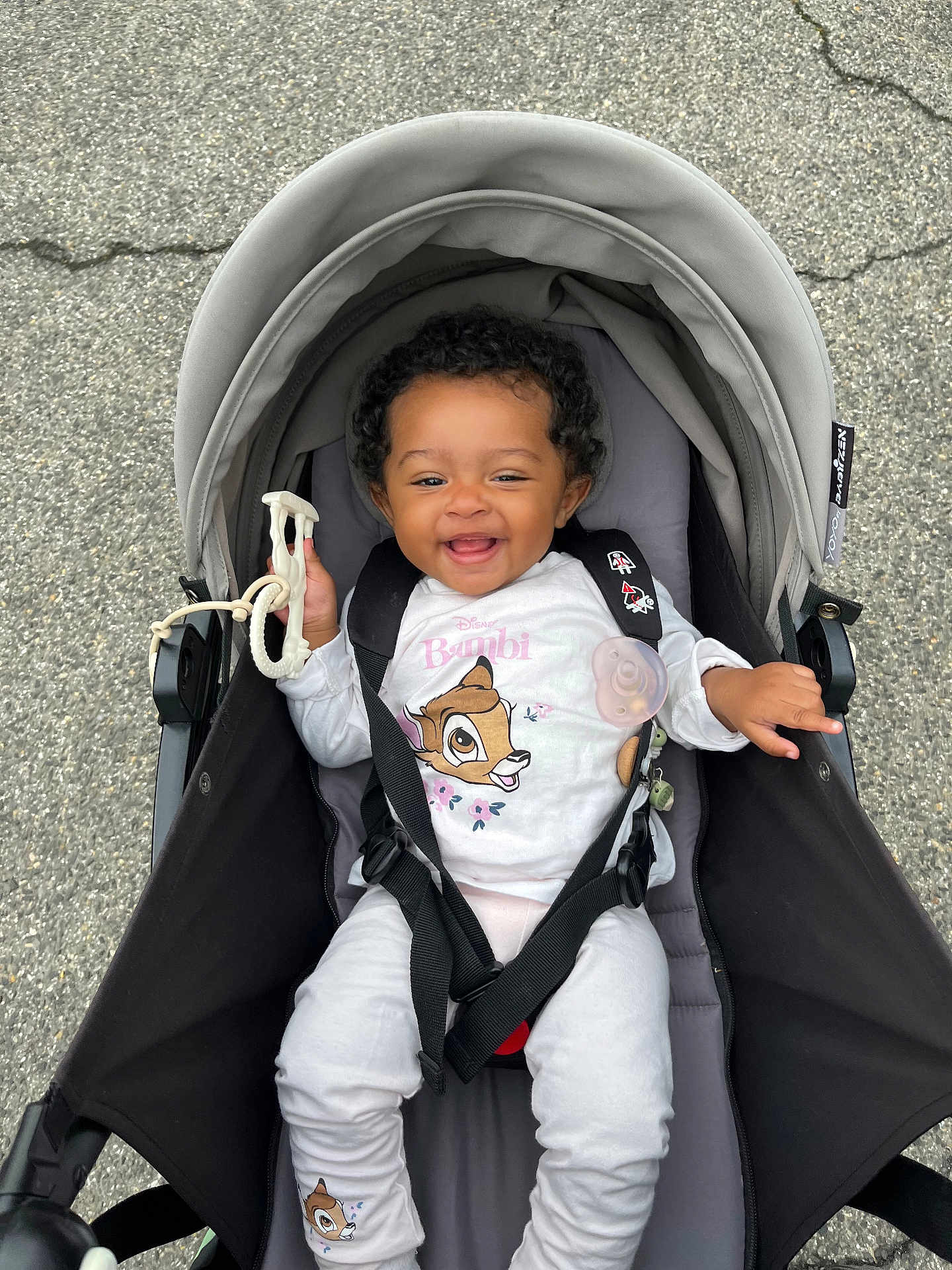 Kayla a rejoint le concours — aidez-le/la à gagner de superbes lots ! baby, stroller, child, smiling, happy, curly_hair, toy, bambi, clothing, outdoor, pavement, seatbelt, infant, cute, sitting, black_straps, hood, person, playful, daylight