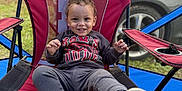 Kash is registered to the contest to win money with this photo: child, toddler, smiling, red_chair, outdoor, camping_chair, sweatshirt, casual_clothing, grass, car, blue_canopy, happy, person, seat, footwear, curly_hair, nature, daylight, relaxed, portrait