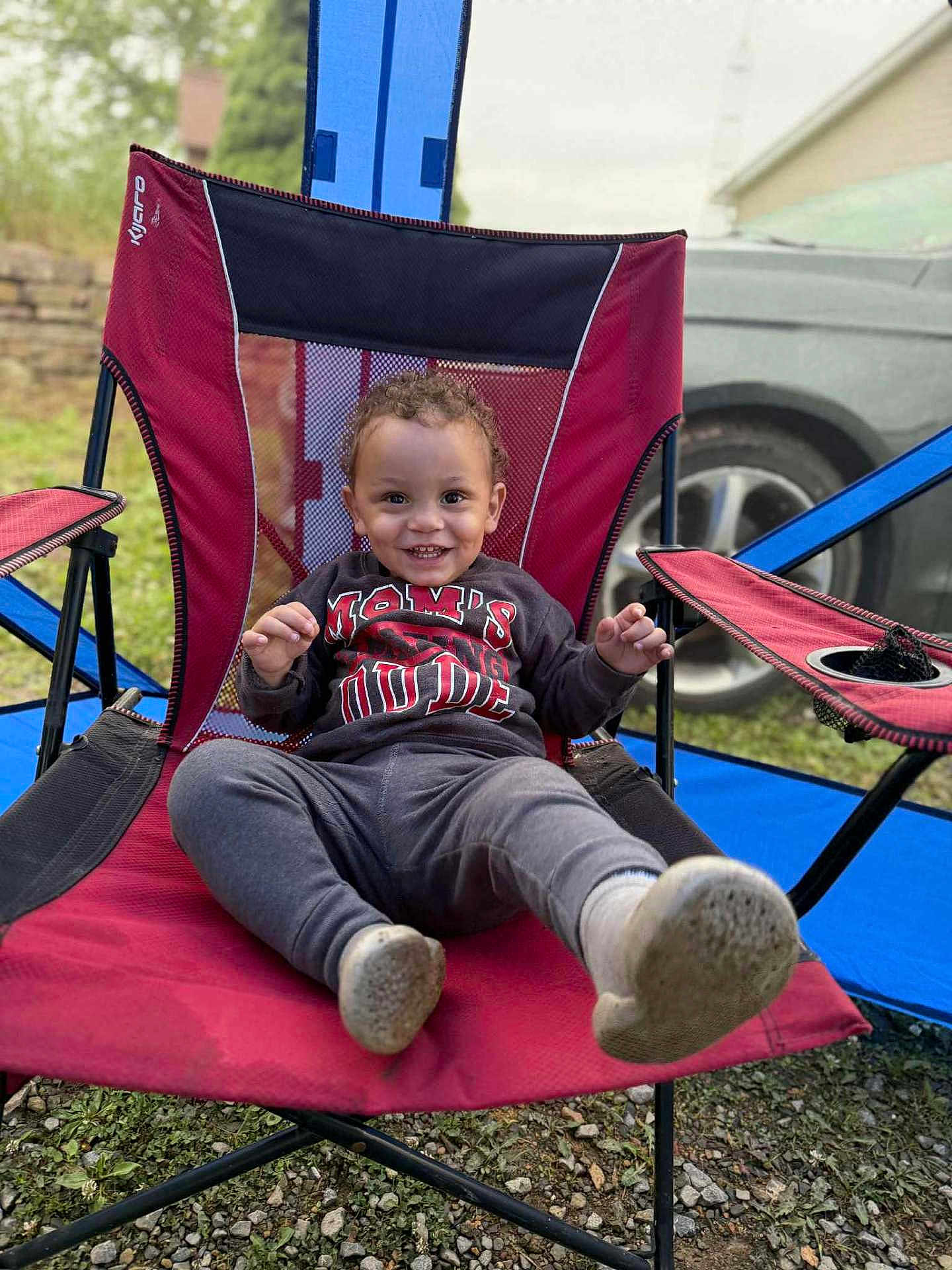 Kash is registered to the contest to win money with this photo: child, toddler, smiling, red_chair, outdoor, camping_chair, sweatshirt, casual_clothing, grass, car, blue_canopy, happy, person, seat, footwear, curly_hair, nature, daylight, relaxed, portrait