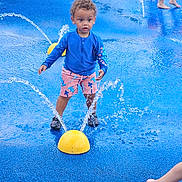 Kash joined the competition — help win amazing prizes! child, toddler, water_play, splash_pad, water_jets, blue_shirt, pink_shorts, yellow_dome, curly_hair, barefoot, outdoor, summer, playground, fun, active, kids, water_splash, recreation, daytime, young_child