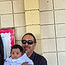 Tommy is registered to the contest to win money with this photo: adult, baby, black_hair, bulletin_board, casual, facial_hair, hand, holding, indoor, infant, light, man, person, portrait, short_hair, smile, sunglasses, tile_wall, wall, white_clothing