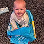baby, child, blue_bag, carpet, smiling, happy, indoor, cute, sitting, recycled_material, playful, person, floor, head, clothing, bright_eyes, rosy_cheeks, expression, fun, home