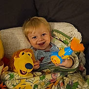 Kole is registered to the contest to win money with this photo: toddler, child, smiling, blanket, plush_toy, lion, toy, couch, indoor, happy, face, person, yellow, blue, orange, flower_pattern, home, cozy, seated, blanket_pattern
