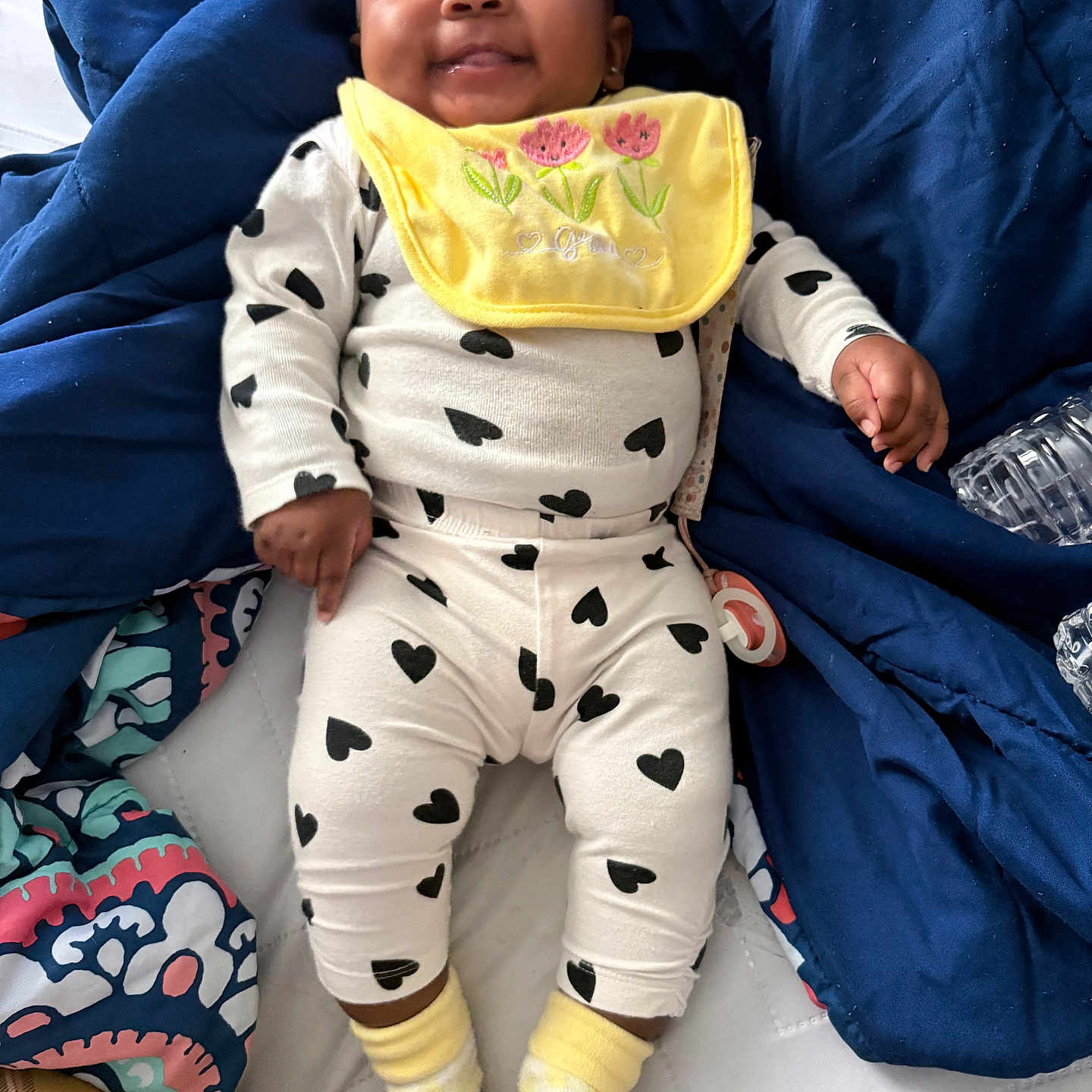 Keior is registered to the contest to win money with this photo: baby, bedspread, black_hearts, blanket, child, cozy, cute, face, happy, indoor, infant, lying_down, pacifier, person, small_hand, smiling, socks, soft_toys, white_outfit, yellow_bib