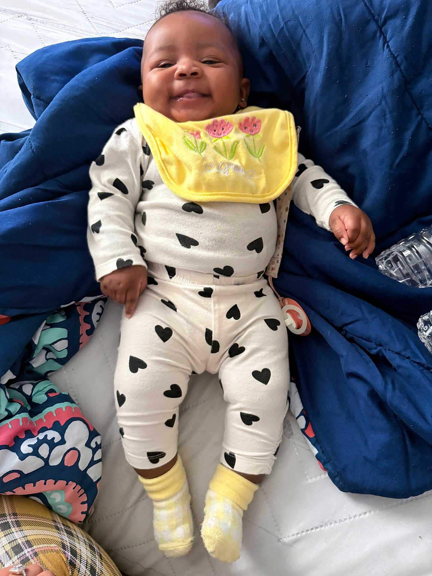 Keior is registered to the contest to win money with this photo: baby, bedspread, black_hearts, blanket, child, cozy, cute, face, happy, indoor, infant, lying_down, pacifier, person, small_hand, smiling, socks, soft_toys, white_outfit, yellow_bib