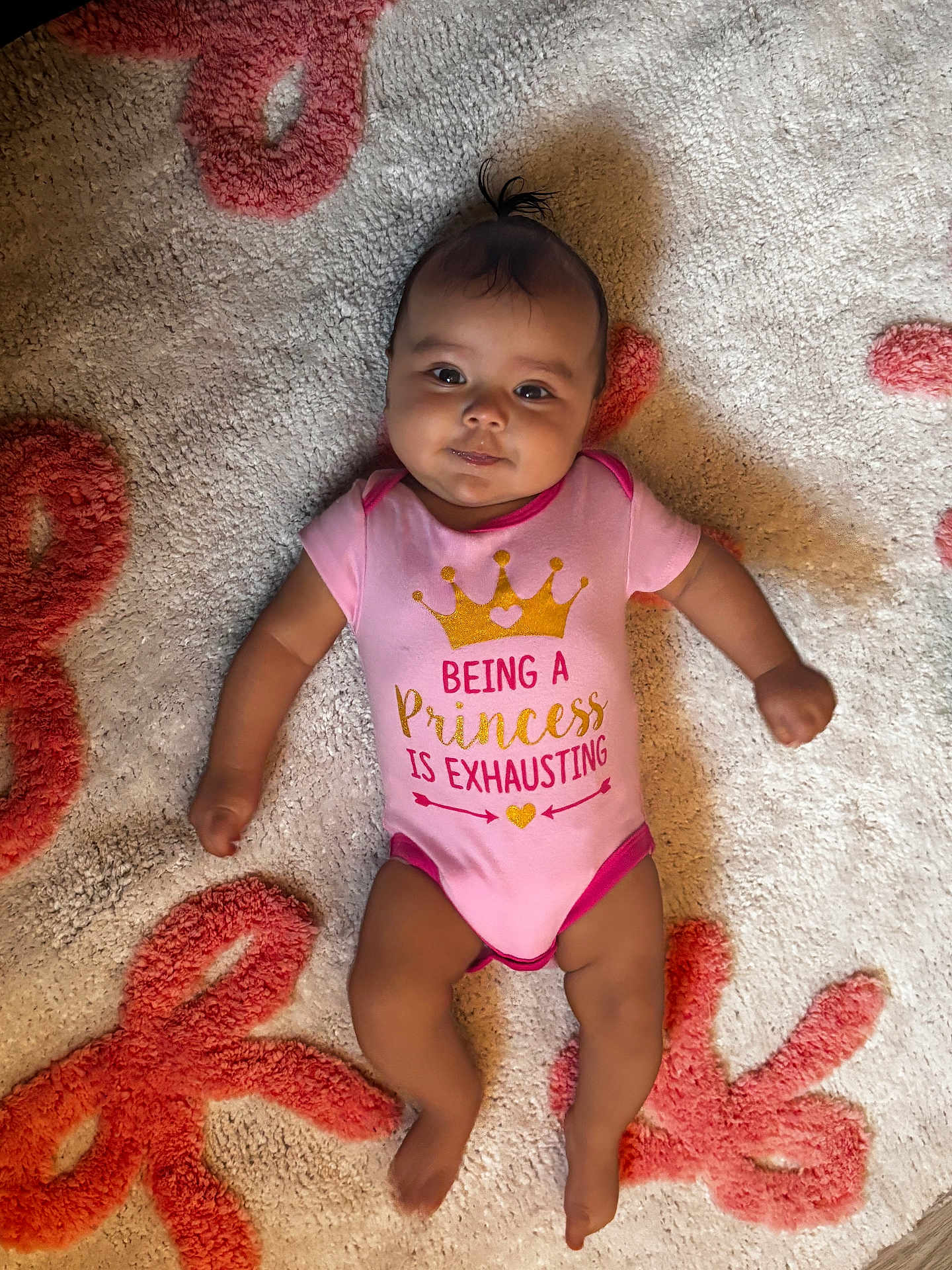 Zaniyah is registered to the contest to win money with this photo: baby, carpet, child, cute, face, floor, happy, indoors, infant, lying_down, onesie, patterned_rug, person, pink_clothing, playful_text, portrait, small_hair_tuft, smiling, soft_texture, young_child