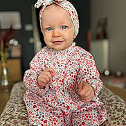 Soline participe au concours pour gagner de l'argent avec cette photo : baby, child, infant, portrait, smiling, blue_eyes, headband, floral_dress, sitting, indoor, cushion, patterned_fabric, cute, young_child, person, clothing, soft_light, closeup, happy, face