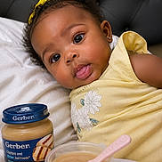 Kehlani is registered to the contest to win money with this photo: baby, infant, girl, yellow_dress, yellow_bow, bed, pillow, baby_food, gerber, spoon, bowl, food, cute, portrait, face, eyes, tongue, skin, head, earrings