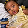 baby, infant, girl, yellow_dress, yellow_bow, bed, pillow, baby_food, gerber, spoon, bowl, food, cute, portrait, face, eyes, tongue, skin, head, earrings