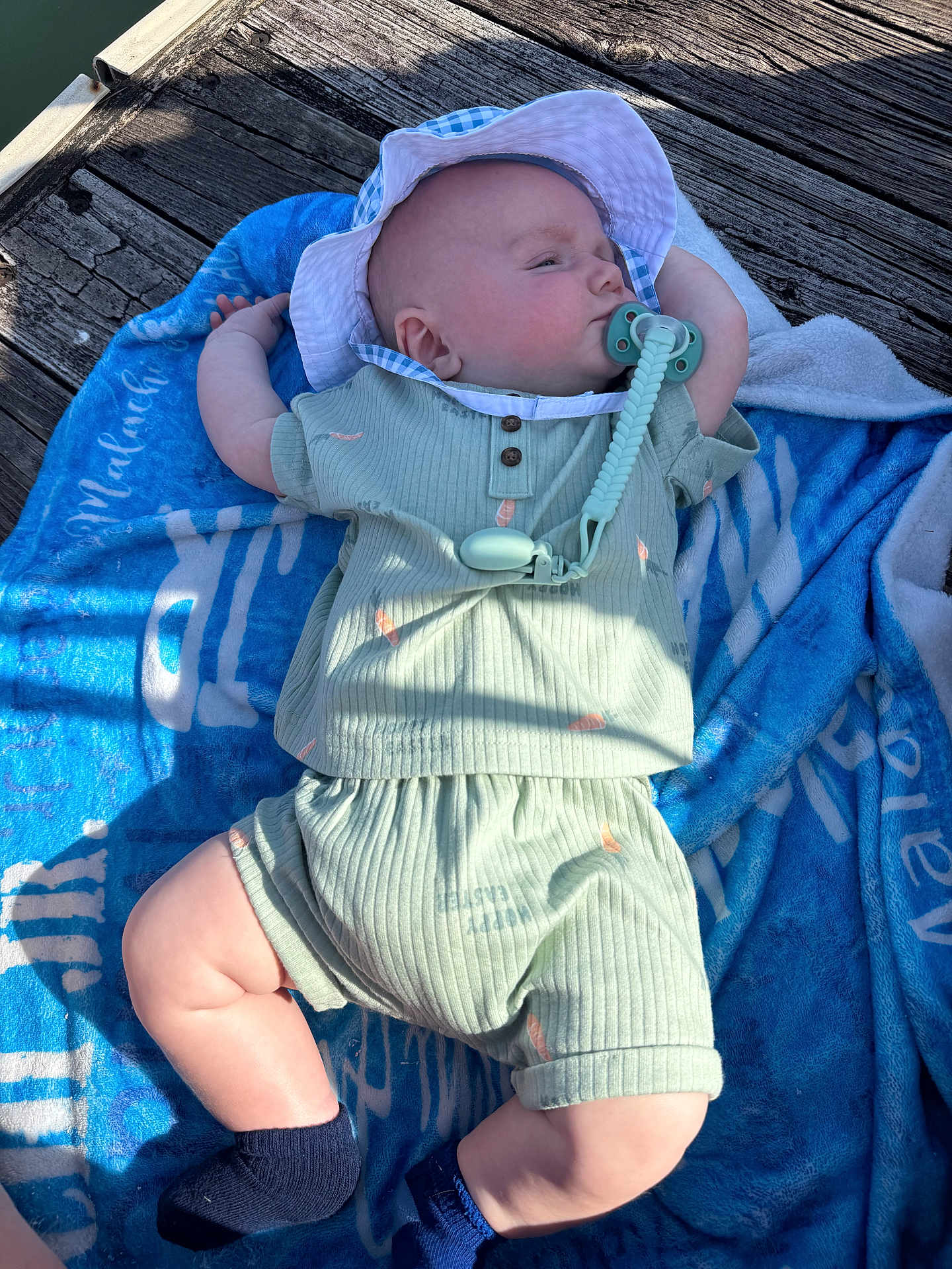 Malachi is registered to the contest to win money with this photo: baby, infant, child, pacifier, sun_hat, hat, towel, wooden_dock, wood_plank, green_outfit, shorts, socks, legs, arm, sunlight, shadow, water, pacifier_clip, relaxed, cute