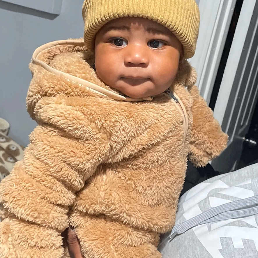 Aayan is registered to the contest to win money with this photo: adult_hand, baby, bear_outfit, child, cute, expression, face, fuzzy_clothing, hand, hat, indoor, looking_at_camera, person, pom_poms, portrait, snackchat_overlay, text_overlay, toddler, warm_clothing, yellow_hat