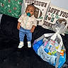 blue, child, couch, decor, face, football, gift_basket, indoor, pacifier, pants, person, pillow, plastic_wrap, ribbon, shirt, sneakers, standing, toddler, toy, white