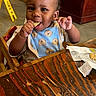Aayan joined the competition — help win amazing prizes! baby, bib, cafe, casual, child, eating, face, food, furniture, happy, highchair, indoor, person, restaurant, seat, smiling, snack, table, toddler, wood
