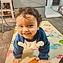 Darvik is registered to the contest to win money with this photo: toddler, child, smiling, crawling, play_mat, indoor, curly_hair, blue_clothing, animal_design, floor, wooden_floor, furniture, cabinet, happy, person, baby, cute, young_child, portrait, home