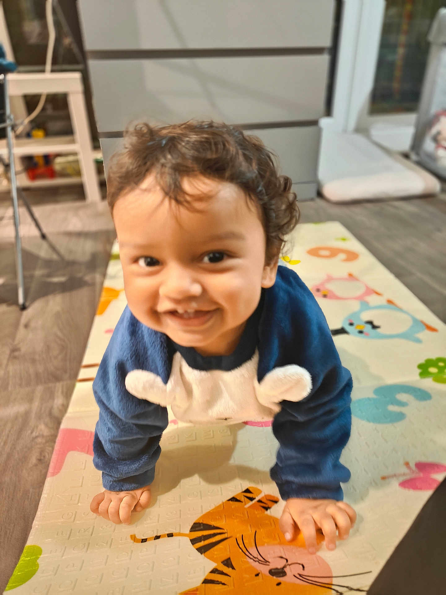 Darvik is registered to the contest to win money with this photo: toddler, child, smiling, crawling, play_mat, indoor, curly_hair, blue_clothing, animal_design, floor, wooden_floor, furniture, cabinet, happy, person, baby, cute, young_child, portrait, home