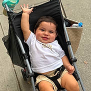Santiago is registered to the contest to win money with this photo: baby, child, stroller, smiling, waving, white_tshirt, beige_shorts, water_bottle, bag, sidewalk, outdoor, happy, cute, person, infant, seat_belt, metal_frame, black_fabric, hand, legs