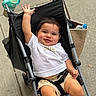 baby, child, stroller, smiling, waving, white_tshirt, beige_shorts, water_bottle, bag, sidewalk, outdoor, happy, cute, person, infant, seat_belt, metal_frame, black_fabric, hand, legs