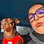 dog, person, glasses, scarf, car, orange_harness, brown_dog, seat, window, daylight, portrait, face, closeup, travel, vehicle_interior, casual_clothing, happy, alert, side_by_side, winter