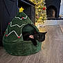 cat, black_cat, pet_bed, christmas_tree_bed, holiday_decor, christmas_tree, stockings, fireplace, flooring, living_room, decorations, plush, cozy, indoor, festive, relaxed, animal, feline, home_interior, ornaments