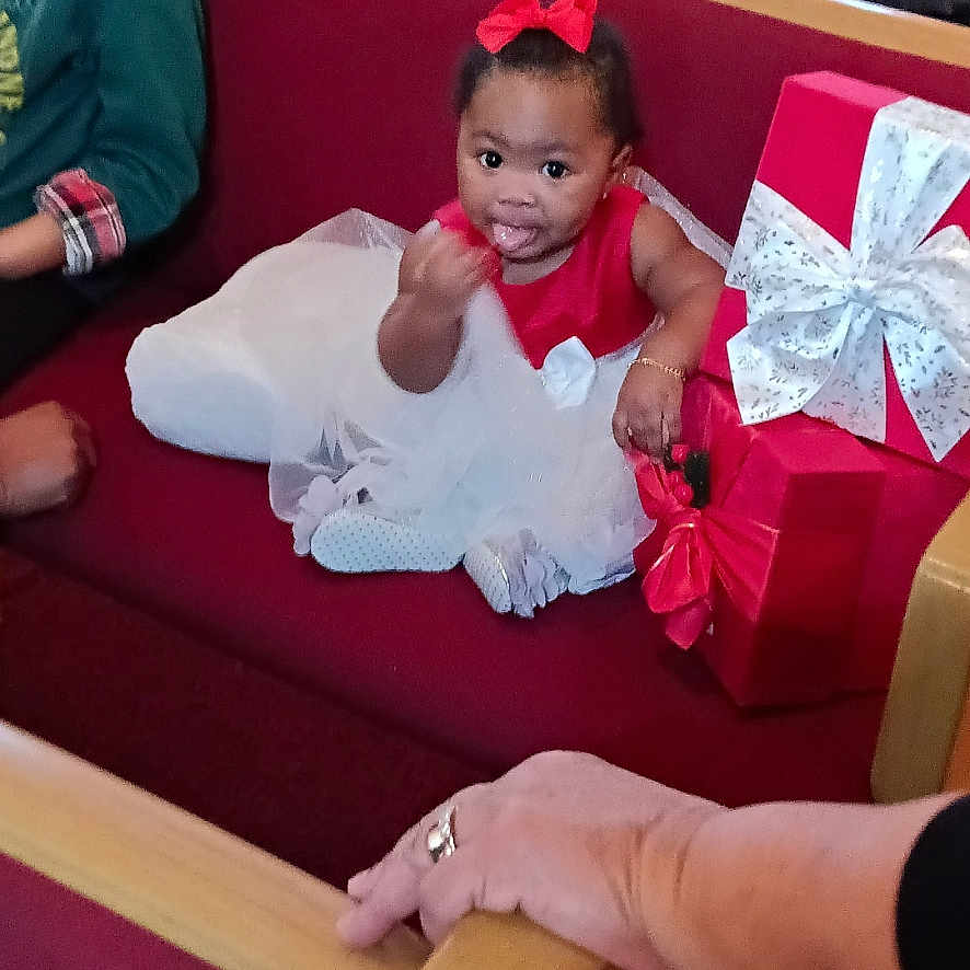 A'Myah joined the competition — help win amazing prizes! adult, bow, celebration, child, church_pew, clothing, festive, gift_box, hand, happy, holiday, indoor, person, pointing, portrait, red_dress, sitting, smile, toddler, wrapped_gift