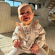 Esmée participe au concours pour gagner de l'argent avec cette photo : baby, infant, smile, sitting, living_room, sofa, rug, sunlight, natural_light, dress, tights, hands, cheeks, portrait, cozy, interior, staircase, window, happy, child